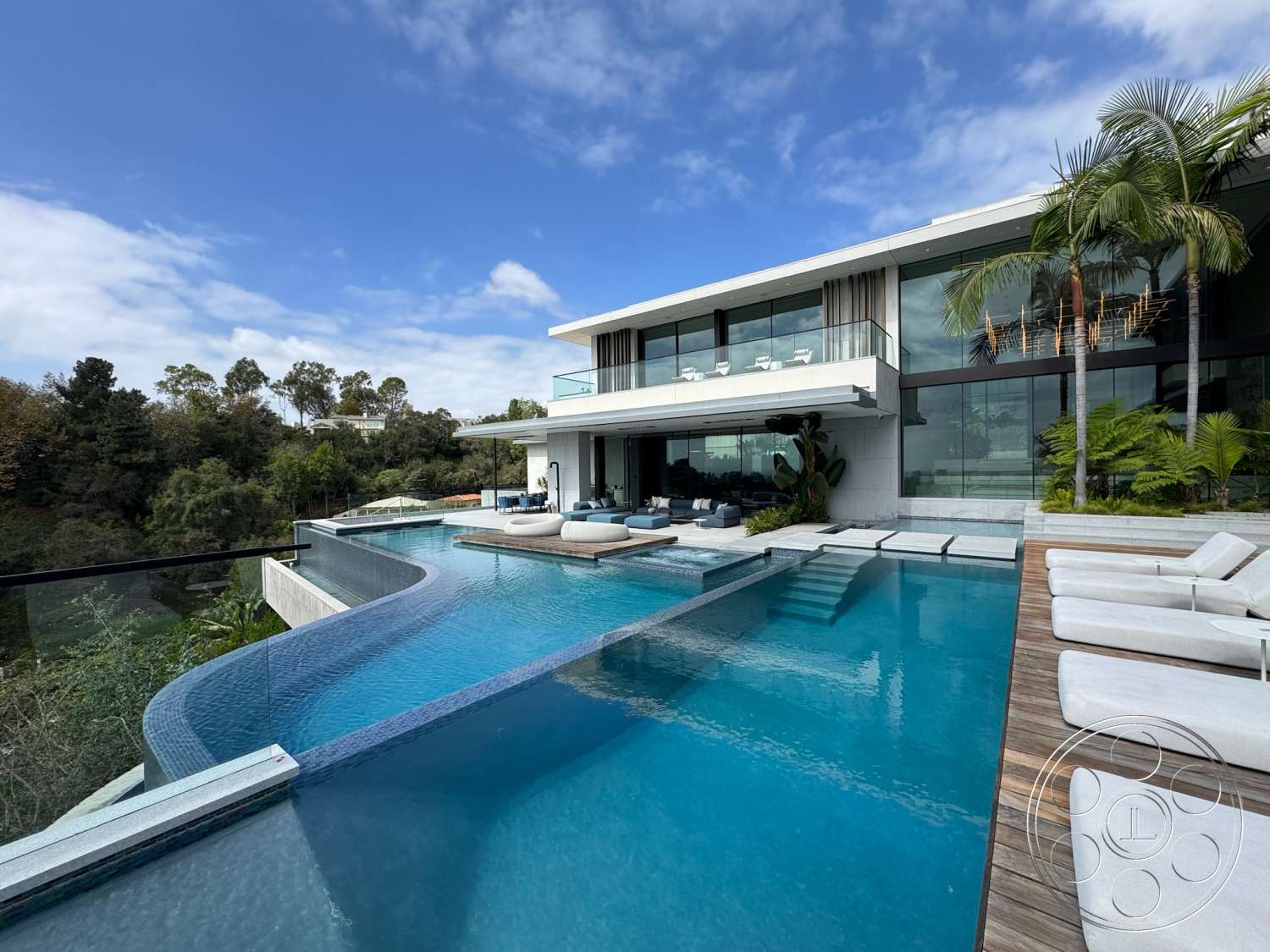 The Bellagio Estate - exterior, outdoor seating area, glass railing, floor-to-ceiling windows, modern architectural style, tropical landscaping, sun loungers, stucco walls, infinity pool, concrete pool deck, wood patio flooring, sloped hillside location, balcony with glass panels