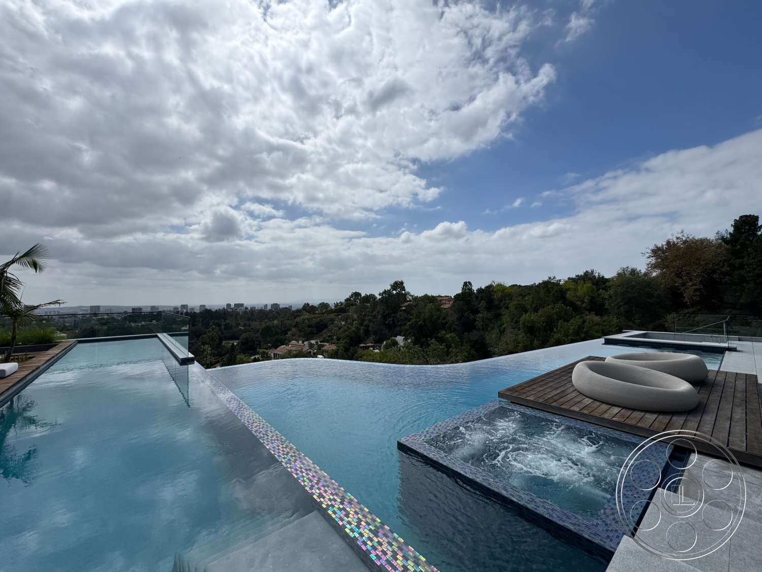 The Bellagio Estate - exterior, glass railing, infinity pool, modern outdoor lounge area, wood deck patio, concrete pool border, panoramic city skyline view, landscaped hillside view, luxury pool area, tile poolside mosaic