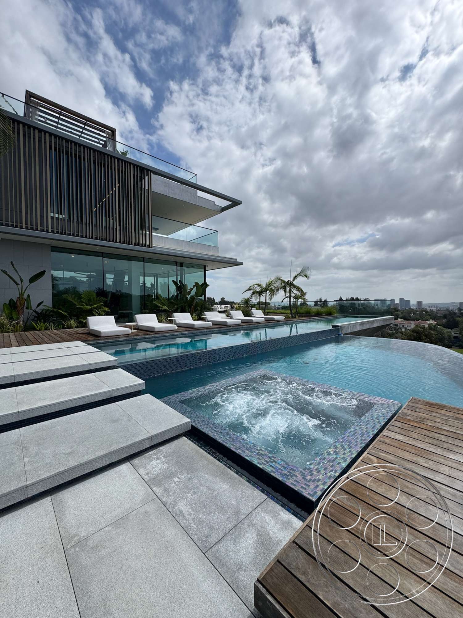 The Bellagio Estate - exterior, glass railings, rectangular swimming pool, floor-to-ceiling windows, lush landscaping, modern architectural style, urban skyline view, infinity pool, concrete pool deck, wood lounge chairs, horizontal wood slat screen, jacuzzi with mosaic tile edge, slab stone steps