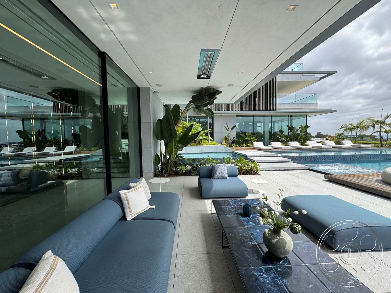 The Bellagio Estate - exterior, contemporary design, glass sliding doors, wooden deck, paved walkways, open outdoor space, landscape lighting, tropical landscaping, infinity pool, rectangular pool, minimalist architecture, stainless steel railing, sliding glass walls, flat roof design, concrete patio flooring, built-in planters, blue outdoor sectional sofa, dark stone coffee table