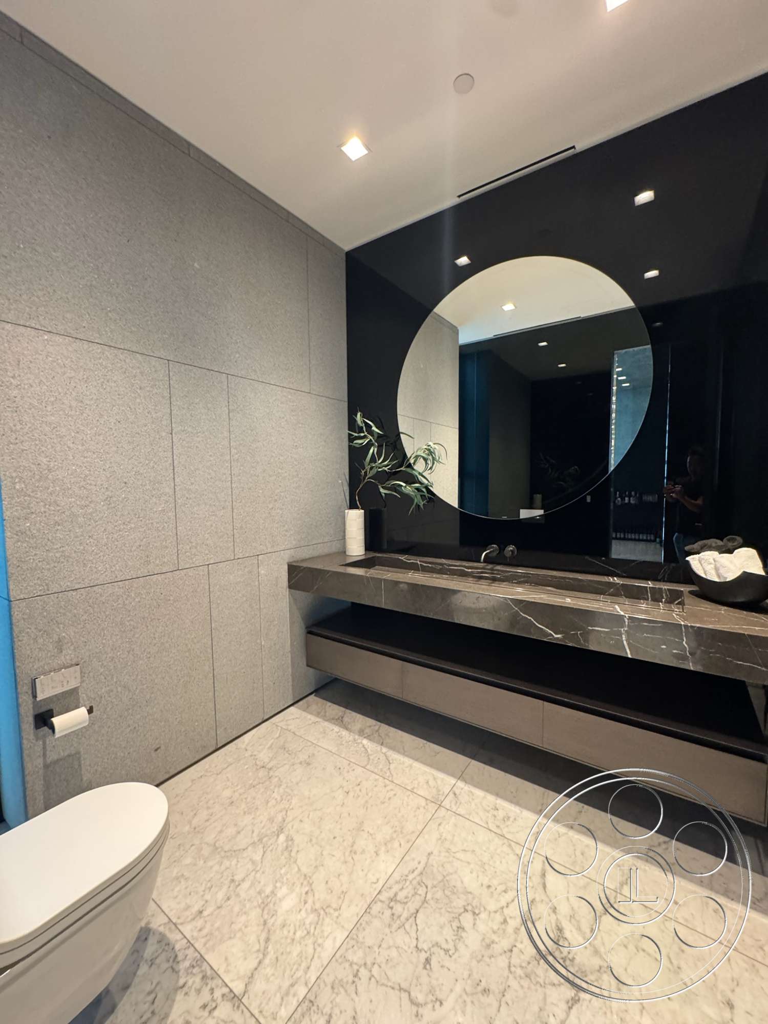 The Bellagio Estate - round mirror, recessed ceiling lights, modern toilet fixture, marble bathroom flooring, grey stone wall tiles, black mirror finish, dark wood floating vanity