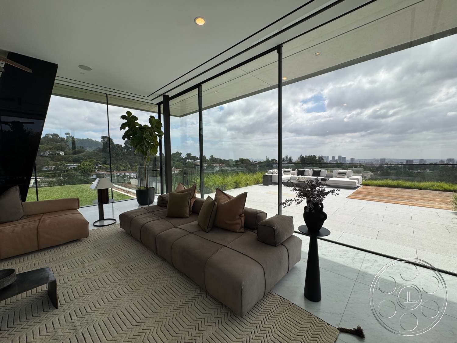 The Bellagio Estate - city skyline view, polished marble flooring, green potted plant, floor-to-ceiling glass walls, natural grass landscaping, open floor plan living room, beige fabric sectional sofa, woven area rug with herringbone pattern, black metal side table, black modern lamp, round black vase, outdoor terrace with lounge seating