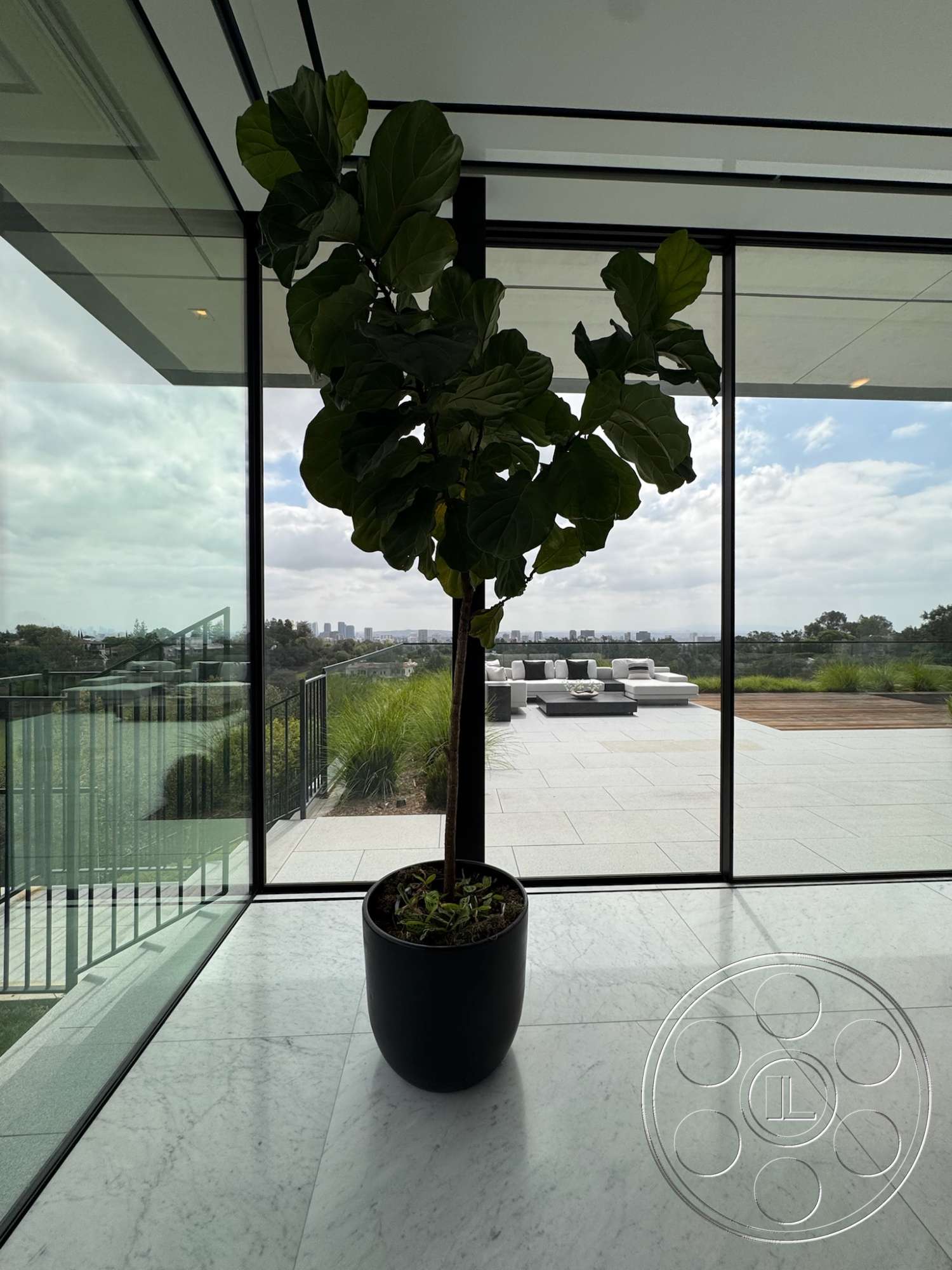 The Bellagio Estate - minimalist design, open space layout, modern architectural style, marble flooring, fiddle leaf fig plant, floor-to-ceiling glass windows, black planter pot, outdoor patio area view, landscape greenery visible