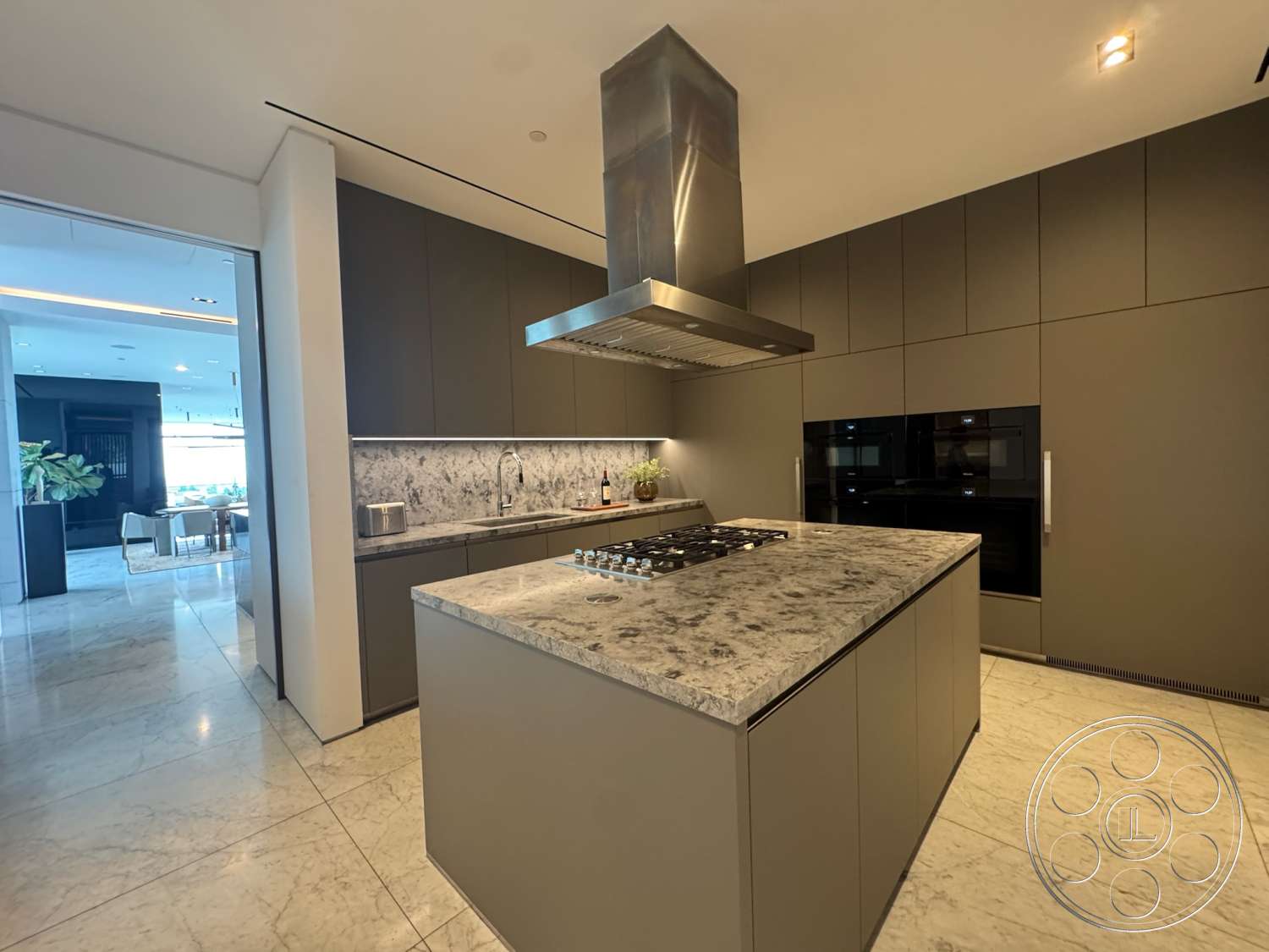 The Bellagio Estate - open floor plan, dark cabinetry, ambient lighting, granite countertop, smooth tile flooring, interior kitchen, stainless steel range hood, marble kitchen island, integrated black kitchen appliances, modern wall-mounted faucet
