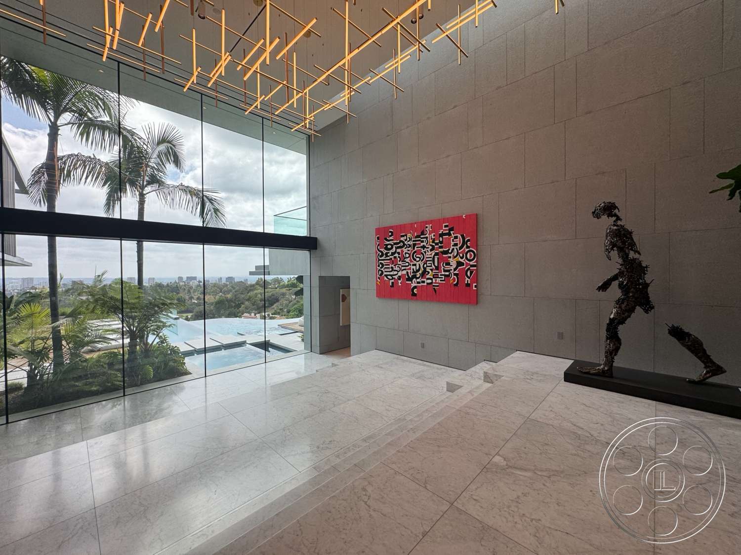The Bellagio Estate - exterior, abstract wall art, floor-to-ceiling windows, lush landscaping, marble flooring, palm trees visible outside, contemporary architectural style, concrete wall panels, modern chandelier lighting, full-height glass windows, open-concept entryway, infinity pool in view, sculptural figure
