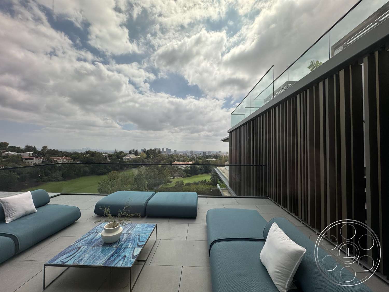 The Bellagio Estate - exterior, modern outdoor furniture, city skyline view, blue fabric sofa, stone patio flooring, glass balcony railing, cloudy sky, decorative potted plant, rectangular patio, wood vertical slat wall, view of green landscape