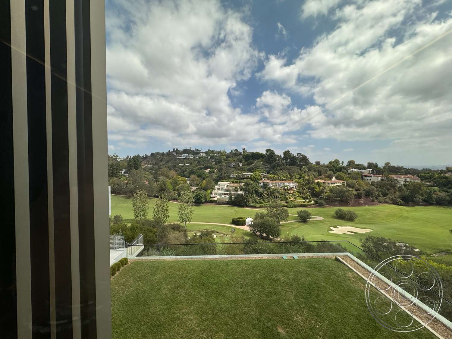 The Bellagio Estate - large windows, exterior, modern architecture, suburban neighborhood, glass railing, concrete patio, green vegetation, landscaped lawn, nature setting, horizontal wooden fence, terraced garden, hilly terrain, cloudy sky, golf course view, two-story homes