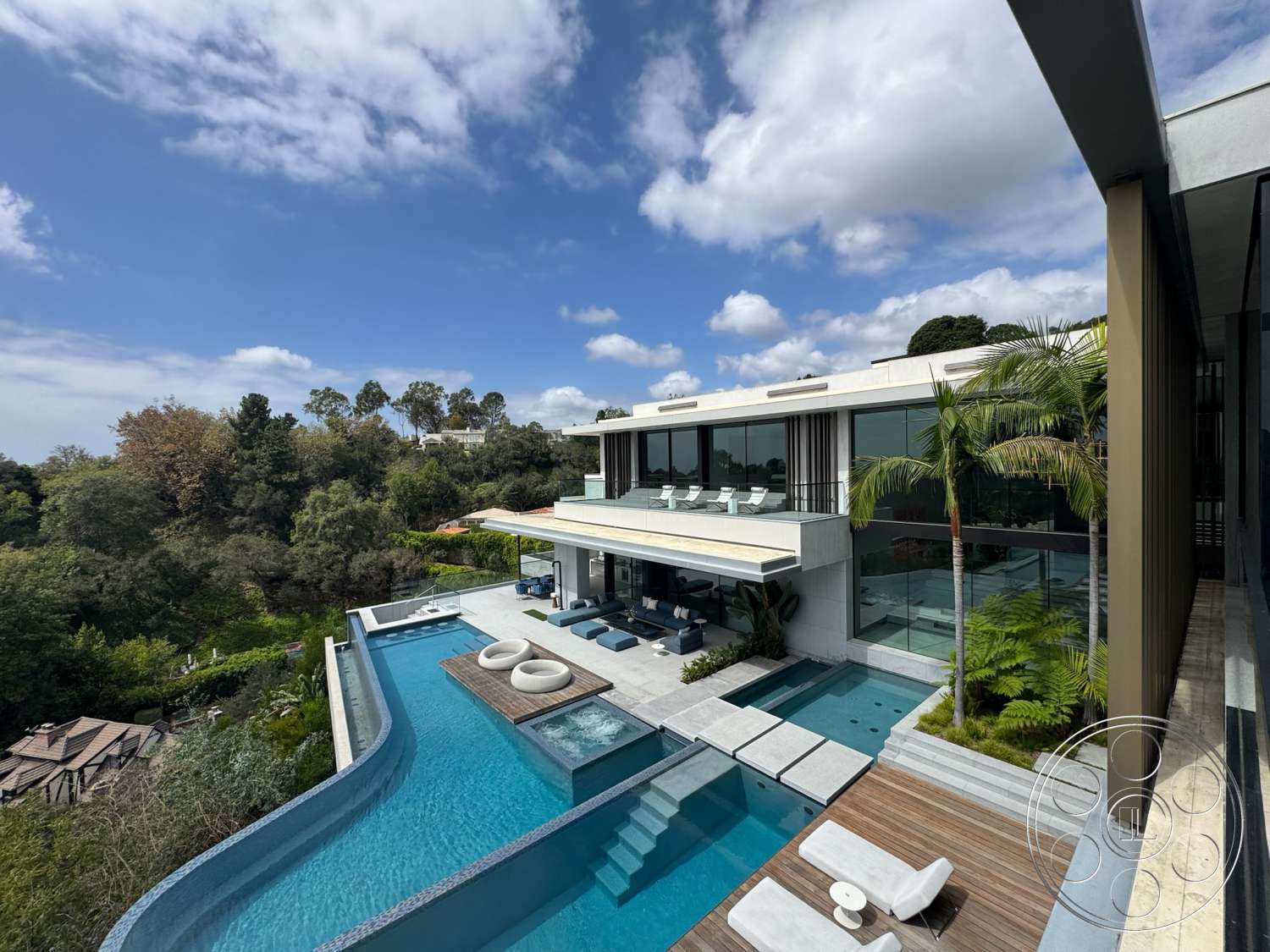 The Bellagio Estate - exterior, exposed concrete walls, sloped terrain, modern architectural style, concrete pool deck, landscaped garden setting, floor-to-ceiling glass windows, stone patio flooring, rectangular infinity pool, palm trees in landscaping, wood deck area, overhanging roof, concrete outdoor steps, glass patio railings, sun loungers on patio