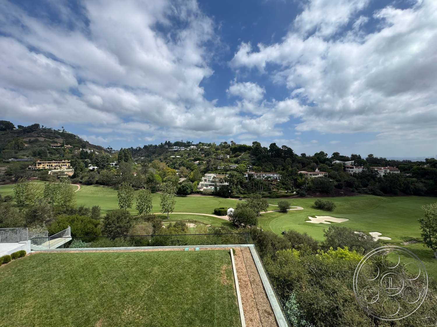 The Bellagio Estate - exterior, contemporary landscaping, natural stone path, rolling hills, glass railing balcony, suburban residential area, lush green lawns, tree-lined hillside, golf course views, granite patio border, scenic mountainous backdrop, overhead clouds