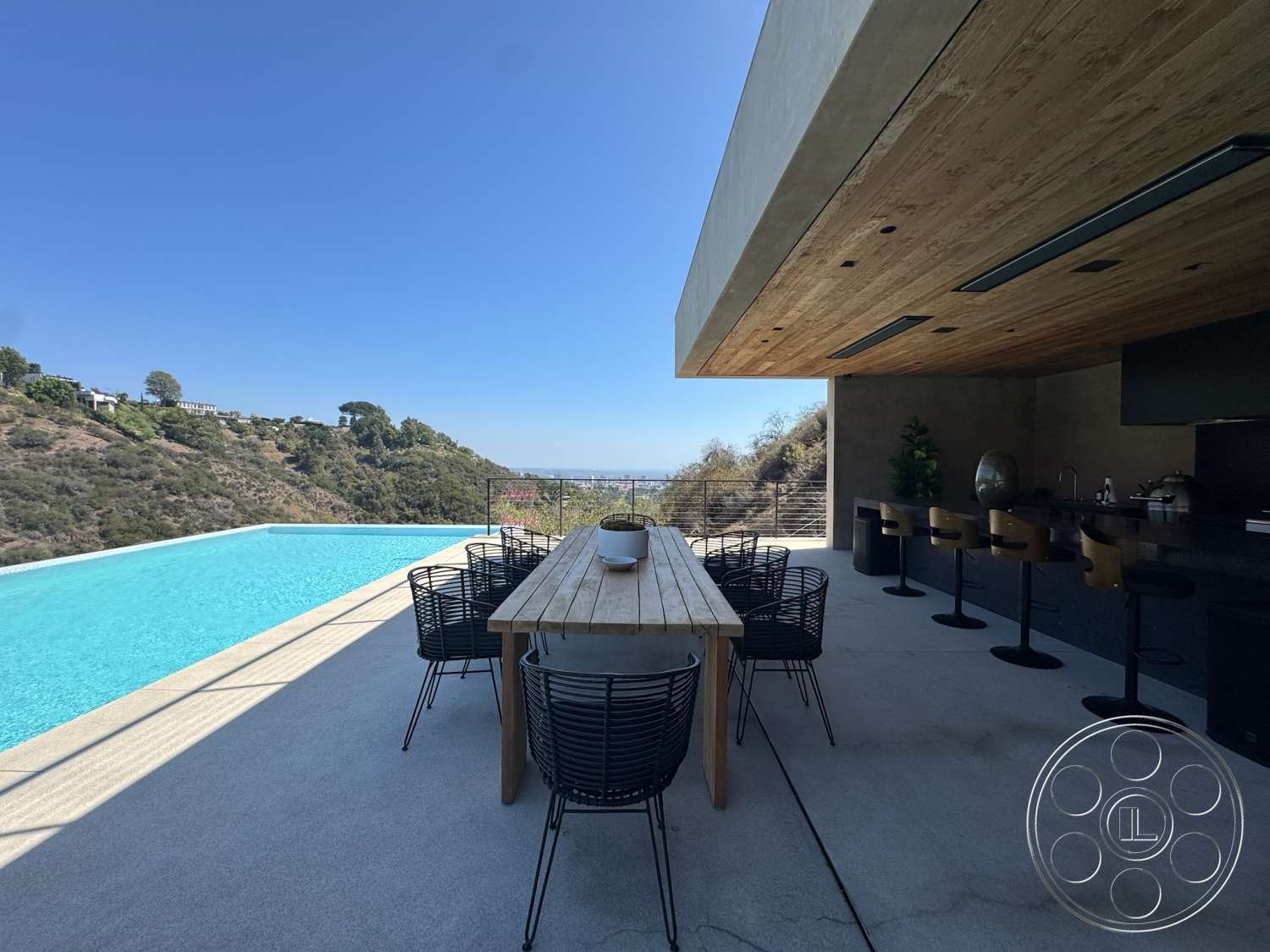 Modern 527 - exterior, wood ceiling, landscape view, infinity pool, glass wall, modern dining table, black metal dining chairs, concrete patio floor, scenic hillside location, sloped hillside, stainless steel outdoor kitchen