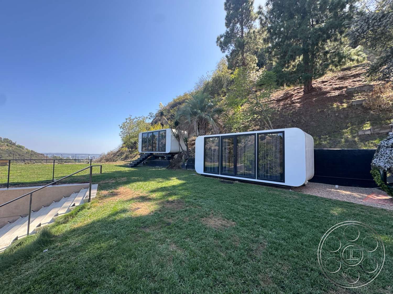 Modern 527 - exterior, large glass windows, surrounding trees, mountain view, sloping hillside, contemporary outdoor living space, wooden deck stairs, modern modular design, white composite siding, landscaped grassy lawn, black privacy wall
