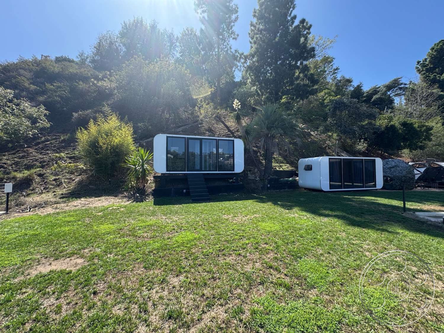 Modern 527 - exterior, green grass lawn, natural hillside setting, large floor-to-ceiling glass windows, wooden steps leading to entrance, white exterior cladding, modern prefabricated structure, black composite deck, rectangular module shape, tropical landscape plants