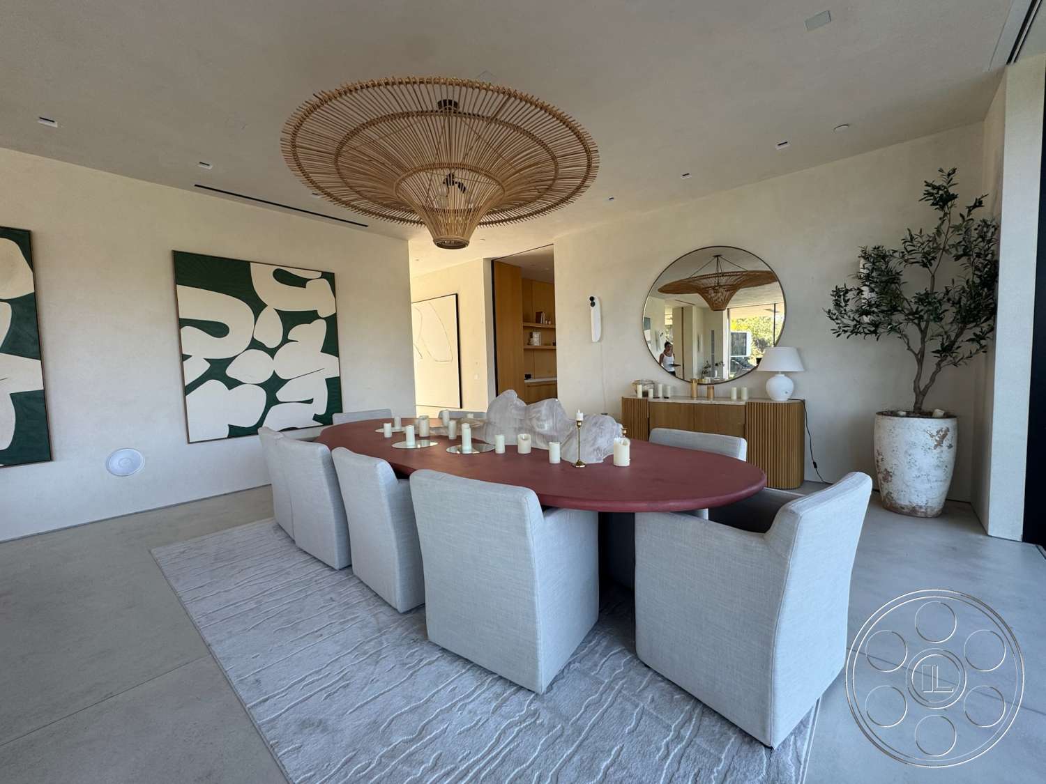 Modern 527 - large windows, concrete flooring, round dining table, neutral area rug, textured plaster walls, woven rattan light fixture, interior dining room, plush upholstered dining chairs, wood sideboard, green abstract wall art, large circular mirror, indoor potted olive tree