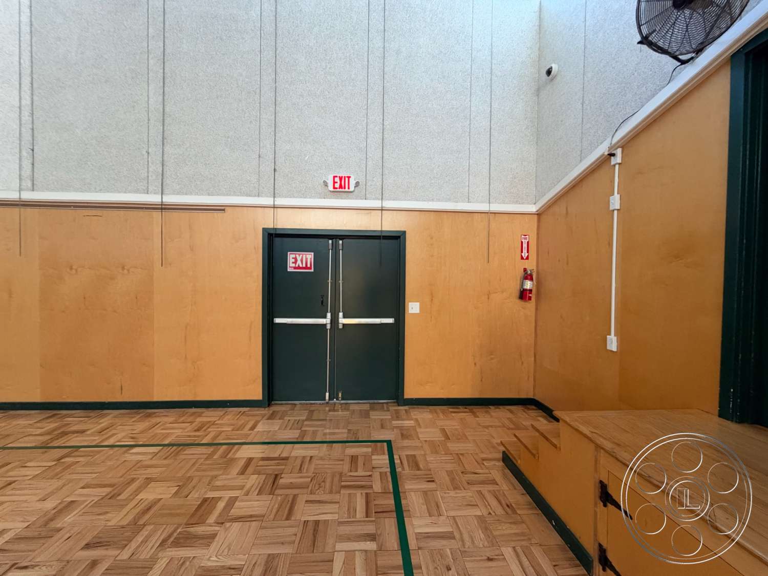 Community Center - parquet hardwood flooring, wooden stage platform, exposed wood paneling walls, fire extinguisher wall mount, green trim details, green exit door, gray acoustic wall paneling, ceiling-mounted fan, parallel hanging wires