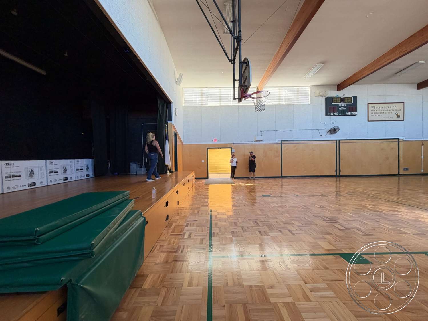Community Center - exposed wooden beams, large windows with natural light, parquet wood flooring, basketball hoop installation, double wooden doors, interior basketball court, light-colored painted walls, vaulted ceiling structure, stage area with curtains, green padded mats, scoreboard wall feature, seating area in gym, gymnasium environment
