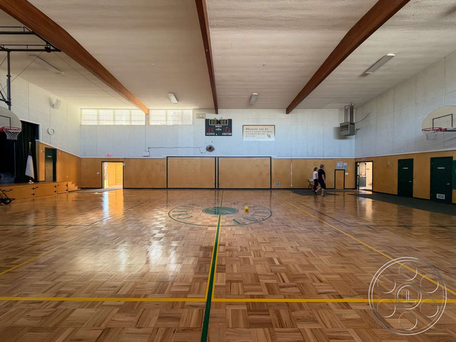 Community Center - large windows for natural light, painted drywall walls, vaulted ceiling with exposed wood beams, interior gymnasium space, parquet wooden gym floor, basketball hoop installations, yellow court markings, green painted doors, wall-mounted scoreboard, gymnasium lighting fixtures