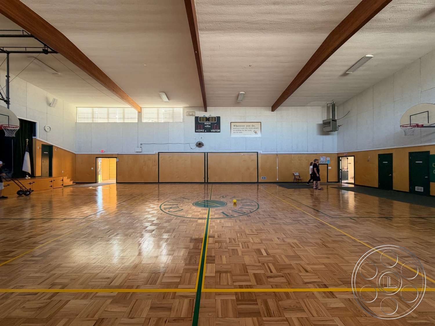 Community Center - floor-to-ceiling windows, textured plaster walls, wood parquet flooring, exposed wood ceiling beams, basketball court markings, wood paneling wall details, interior gymnasium, green painted doors, wall-mounted scoreboard, yellow painted court lines, backboard and hoop structures, seating steps for spectators, automatic air conditioning unit, open ceiling with light fixtures