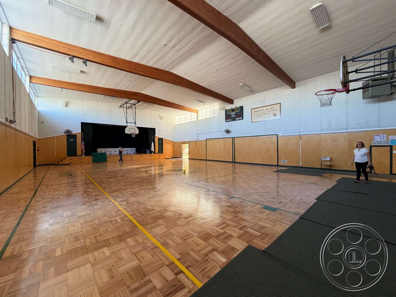 Community Center - floor-to-ceiling windows, glass windows, basketball hoop, stage area, ceiling-mounted lighting, wood beam ceiling, white painted walls, exposed wooden ceiling beams, green accent walls, gymnasium hardwood flooring, parquet wood floor, padded gym mats, basketball court lines, scoreboard wall