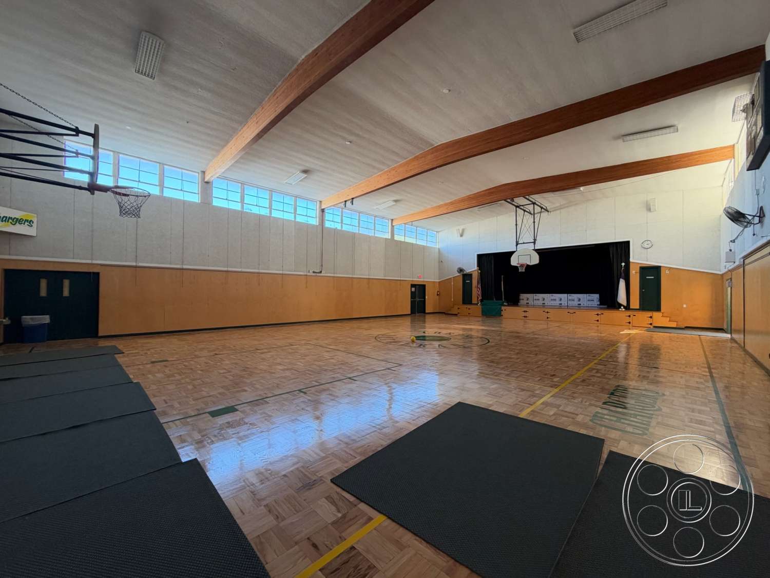 Community Center - wooden ceiling beams, auditorium style seating, exposed ceiling, basketball hoop, exercise mats, wooden wall panels, acoustic wall panels, large classroom windows, green double doors, gymnasium lighting fixtures, gymnasium interior, high ceiling gymnasium, parquet basketball court floor, theatrical stage area, yellow court line markings, gymnasium wall mount fans