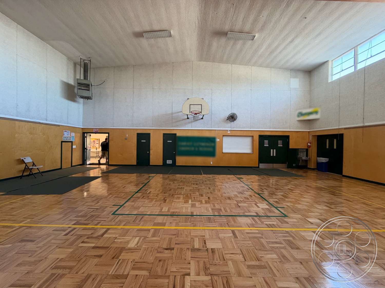 Community Center - basketball hoop, wall-mounted fans, parquet wood flooring, white wall panels, interior gymnasium space, vaulted ceiling with acoustic panels, green painted doors, hardwood floor basketball court, illuminated by fluorescent ceiling lights, black exercise mats