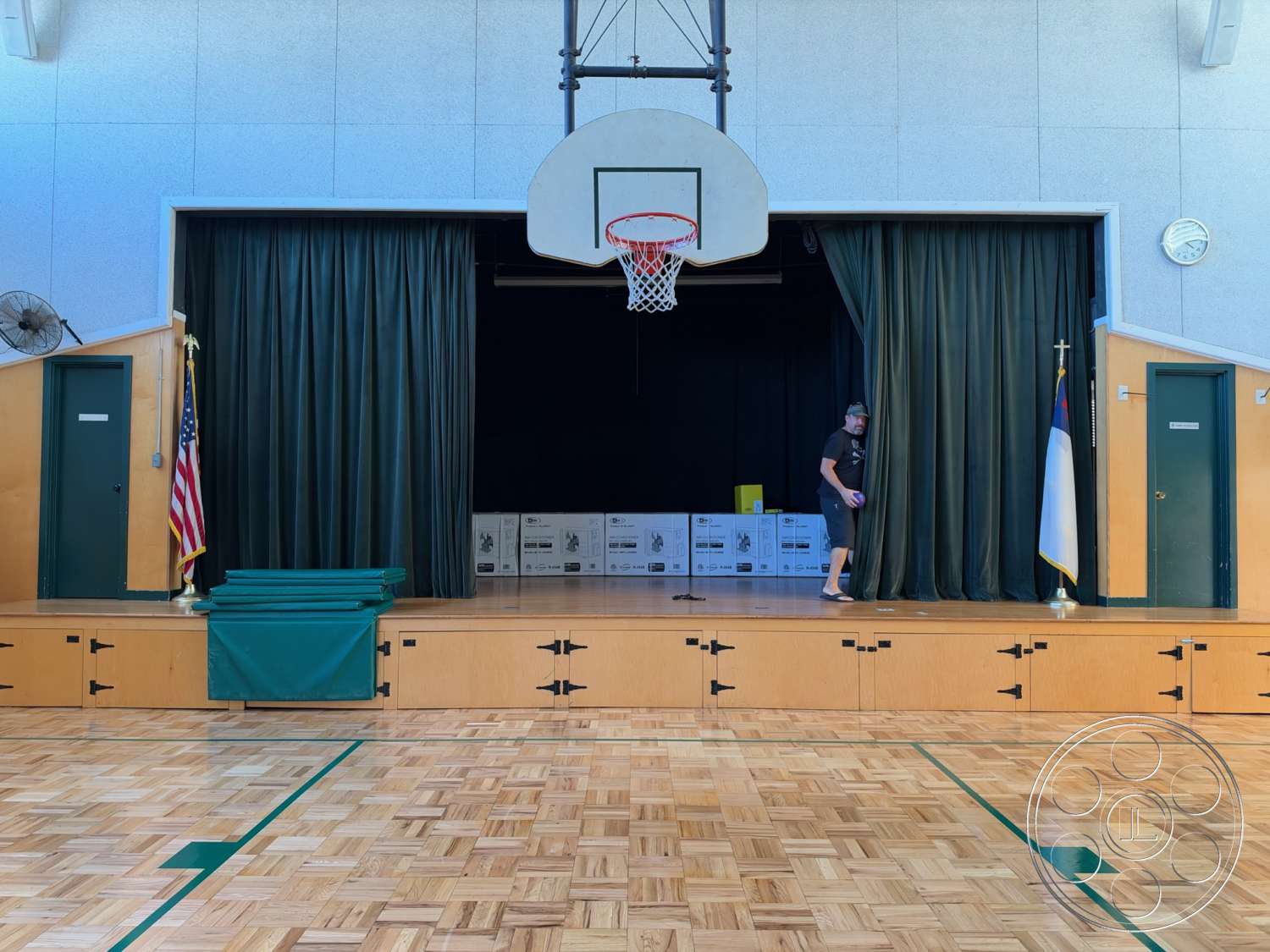 Community Center - draped curtains, wood parquet flooring, crown molding details, wooden stage platform, American flag display, clock mounted on wall, interior gymnasium, green curtain stage backdrop, basketball hoop mounted on wall, exposed ceiling with lights, green folding mats, dark green doors, stacked boxes in backstage area