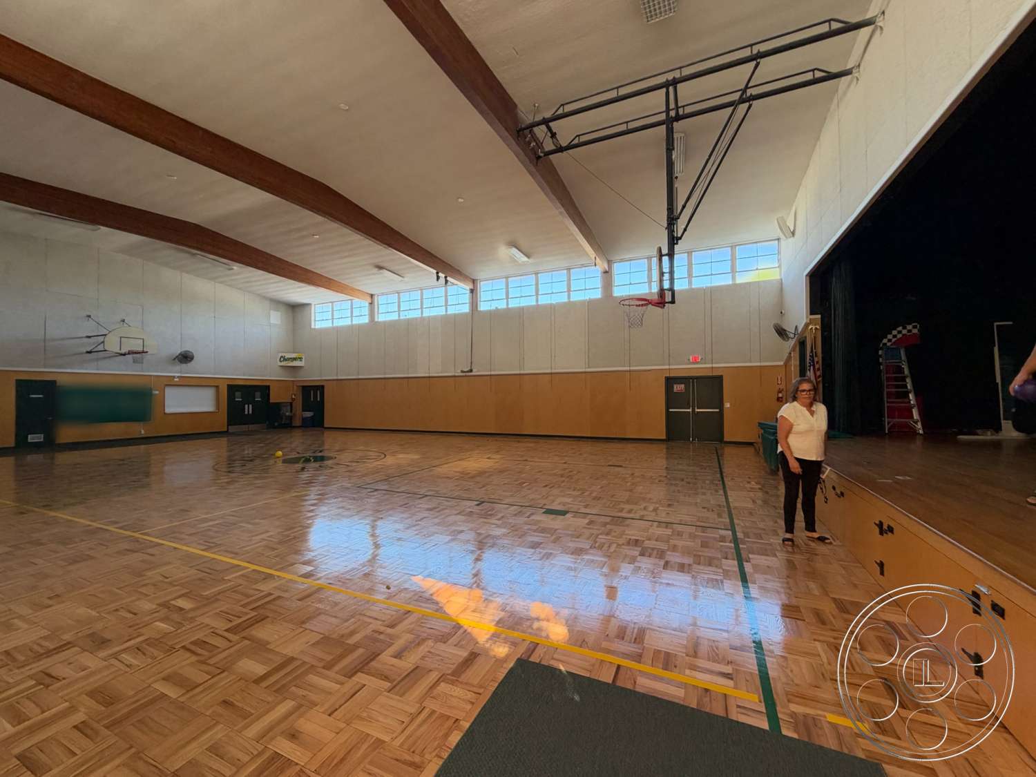Community Center - exposed concrete walls, large open space, basketball hoop, performance area, stage area, wood beam ceiling, emergency exit doors, bright overhead lighting, gymnasium hardwood flooring, high windows, wood panel wainscoting, speaker system, track markings on floor, thick stage platform
