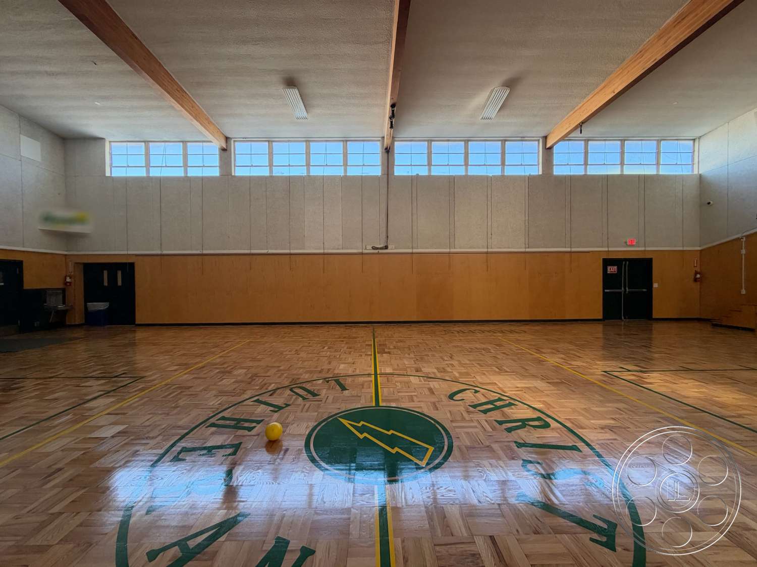 Community Center - floor-to-ceiling windows, exit sign, fire extinguisher, acoustic ceiling tiles, exposed wood ceiling beams, parquet wood flooring, wood paneling walls, basketball court markings, interior gymnasium, green accent lines, yellow rubber ball