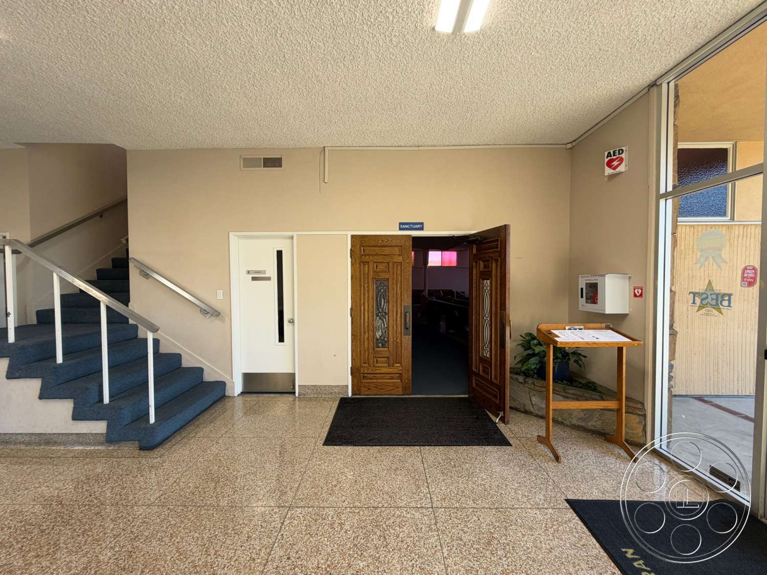 Community Center - ceramic tile flooring, exposed concrete wall, glass entrance door, interior entrance hall, metal stair railings, natural lighting from windows, architectural signage, blue carpeted staircase, decorative wood double doors, rubber mat floor runner
