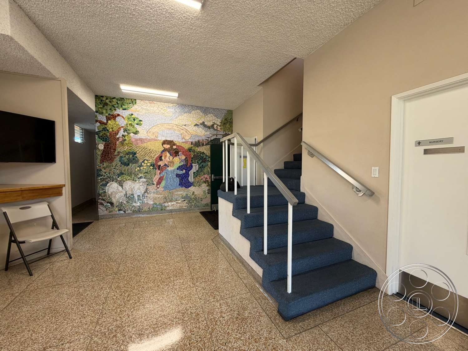Community Center - exterior, floor-to-ceiling windows, wooden accent wall, textured ceiling panels, granite floor tiles, white painted wall, open foyer space, light fixture ceiling, blue carpeted staircase, mosaic wall mural, glass paneled railing