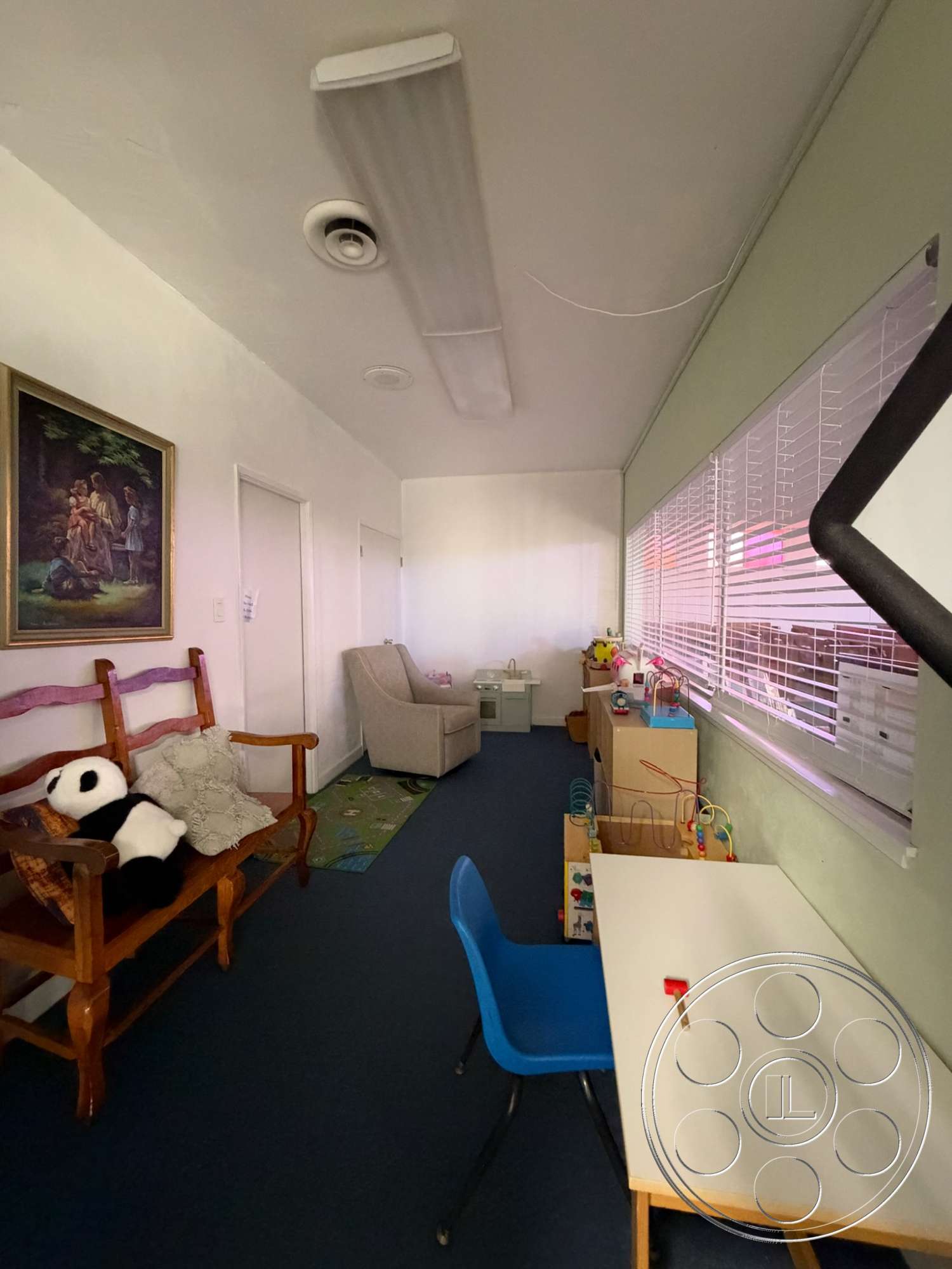 Community Center - framed artwork, carpet flooring, plush armchair, painted drywall walls, wooden bench seat with cushions, blue plastic chairs, blinds window treatment, white laminate desk, children's activity area, light ceiling fixtures, playroom setting