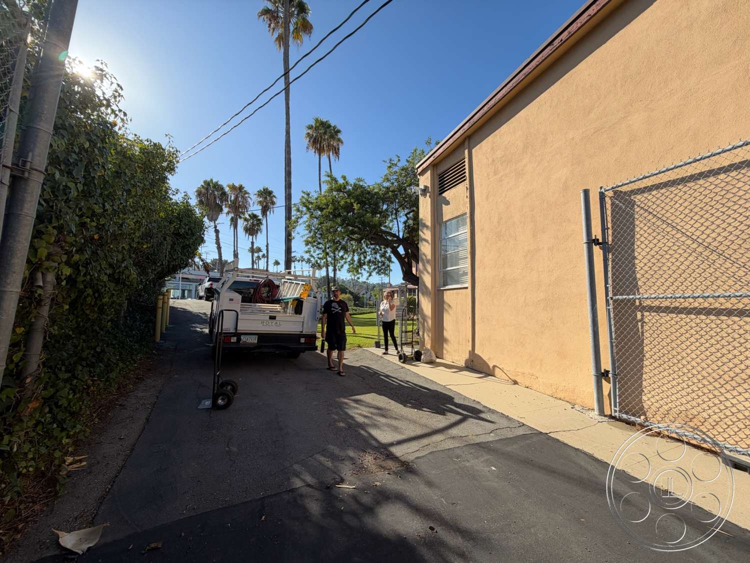 Community Center - exterior, clear blue sky, concrete sidewalk, palm trees in background, metal chain-link fence, stucco building wall, asphalt alleyway, utility truck