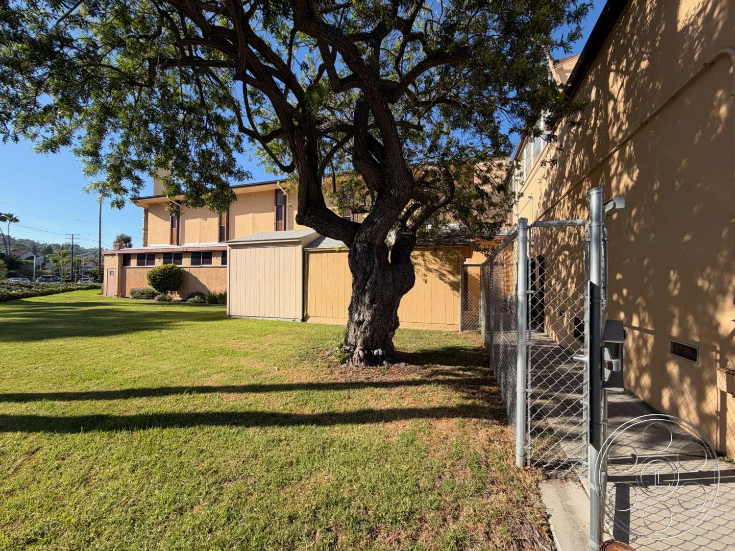 Community Center - exterior, landscaped garden, grass lawn, wooden shed, chain-link fence, outdoor utility area, single-story building, mature tree shade, painted stucco walls
