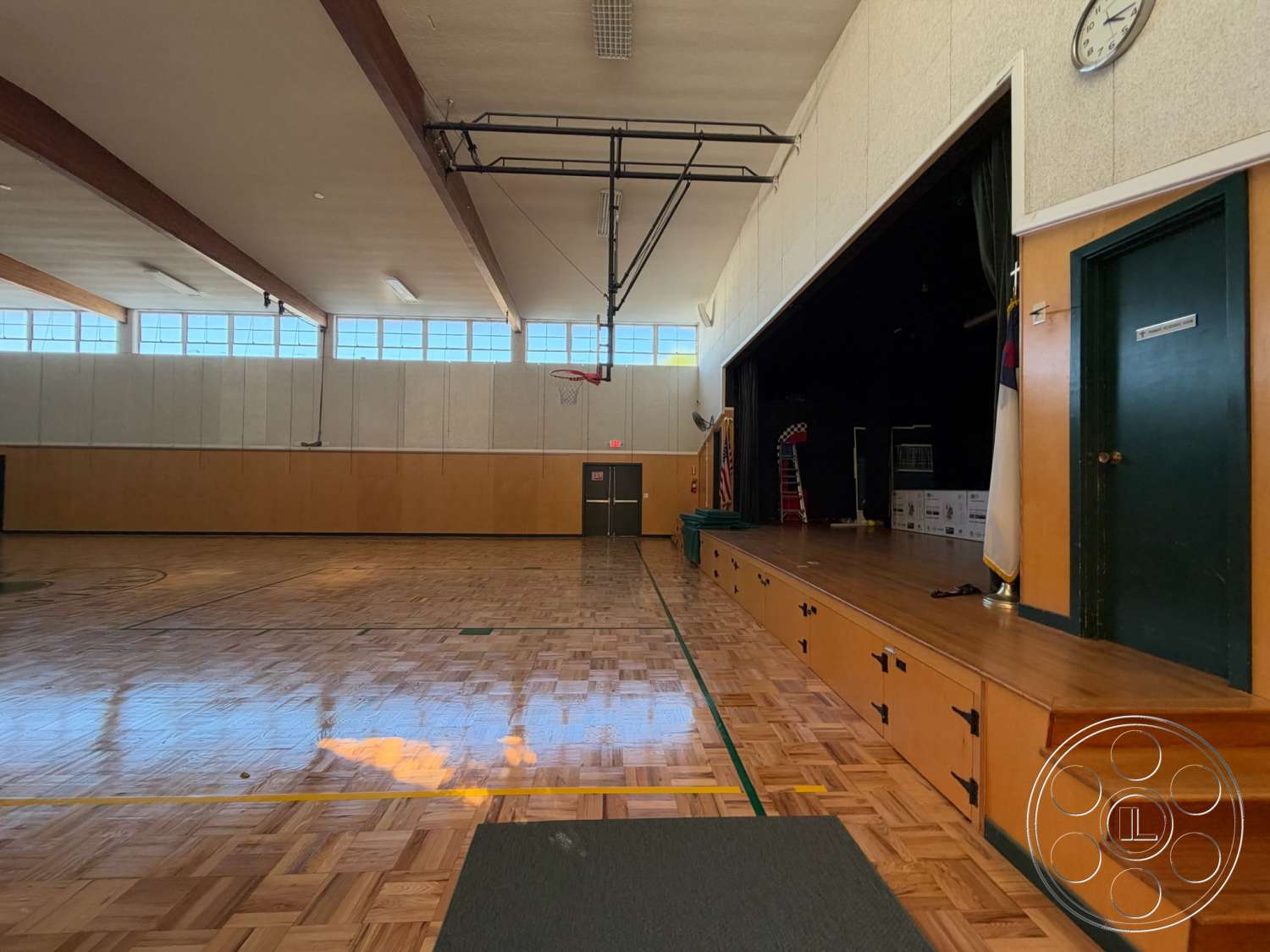 Community Center - large windows, green accent wall, exposed wooden ceiling beams, clock mounted on wall, interior gymnasium, wooden gym floor, stage area with wooden platform, indoor basketball court with markings, tile wall paneling, ceiling-mounted basketball hoop, dual door entrance, wooden stairs leading to stage