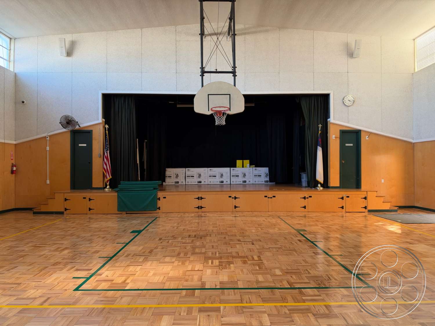 Community Center - exposed wood panel walls, American flag display, fire extinguisher wall mount, high ceiling with integrated lighting, stage area with black curtain, basketball hoop with backboard, wood parquet gym floor, green painted wooden doors, green gym line markings, auditorium-style stage, wooden steps leading to stage