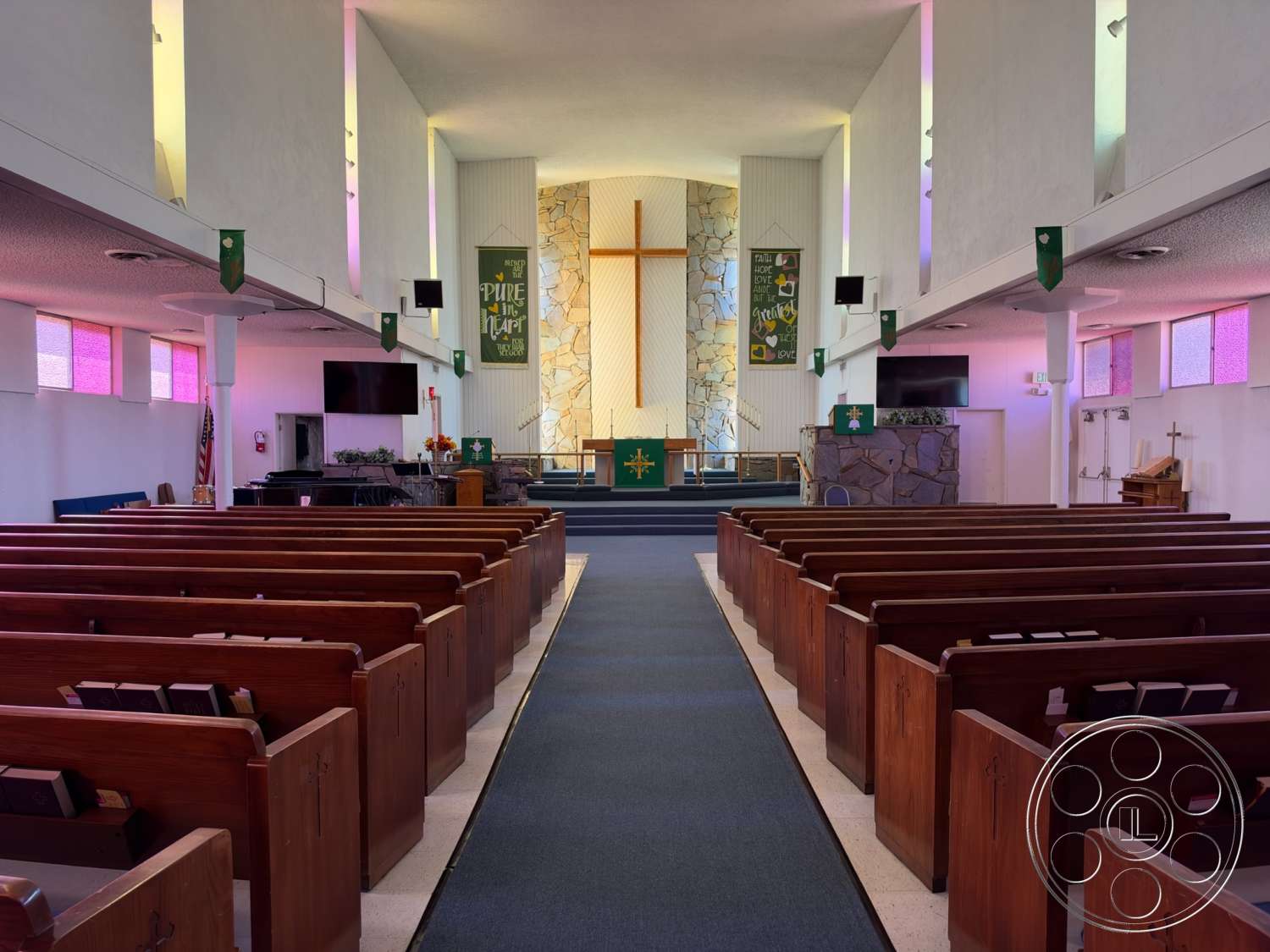 Community Center - natural stone accents, open floor plan, suspended lighting fixtures, vaulted ceiling, wooden pews, walls with textured plaster finish, carpeted aisle flooring, stone feature wall behind altar, colored glass windows, built-in wooden altar, liturgical banners, media display screens, raised platform for altar