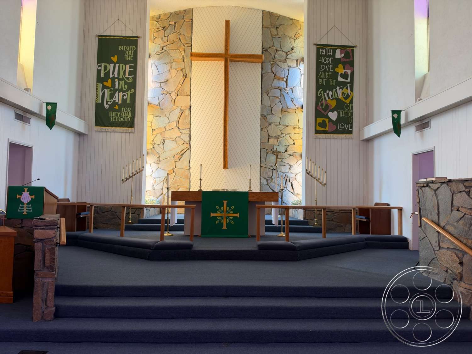 Community Center - floor-to-ceiling windows, carpet flooring, vaulted ceiling, built-in bookshelf, exterior stone feature wall, wood paneling walls, raised platform stage, cross architectural detail, altar with wooden construction, interior sanctuary, decorative banners, multi-level seating areas, lit candle holders
