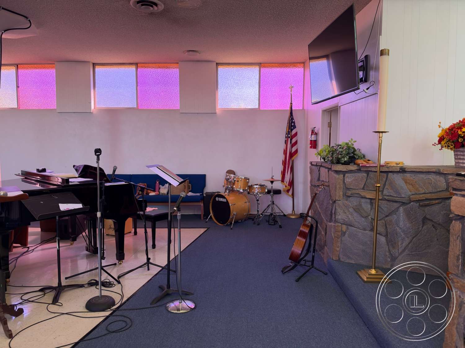 Community Center - carpet flooring, American flag, stone fireplace wall, indoor potted plants, textured ceiling tiles, wooden piano, blue upholstered seating, mounted flat-screen television, colored glass windows, metal microphone stand, wooden drum set, traditional altar candles, wooden acoustic guitar