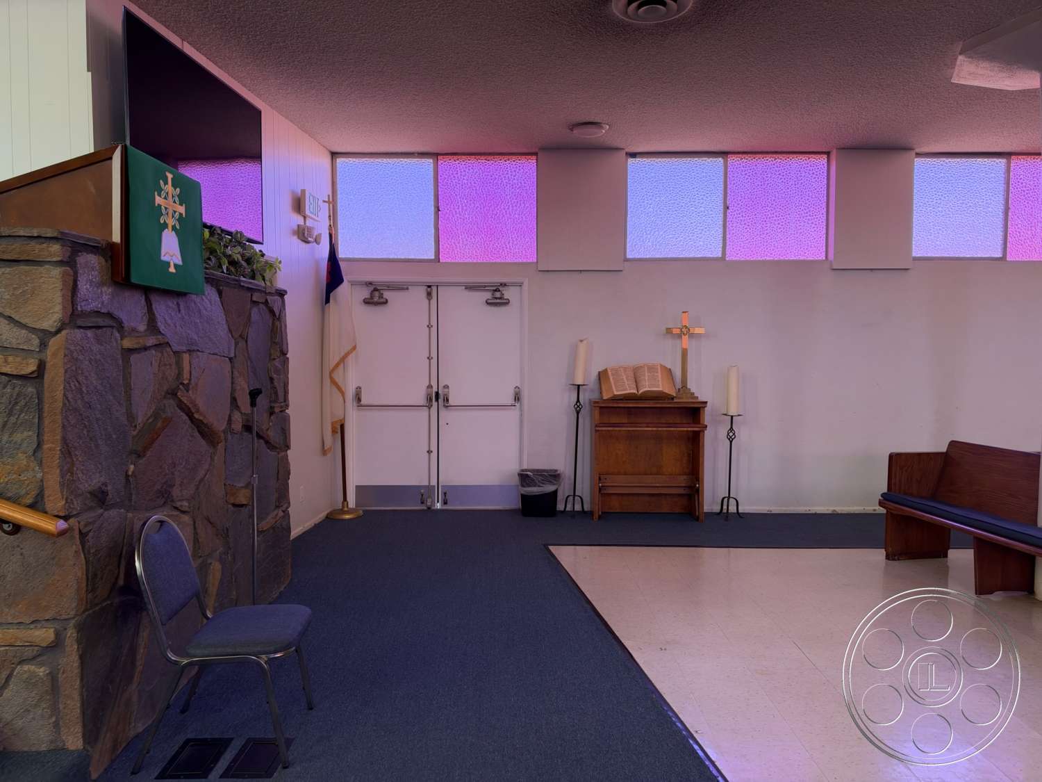 Community Center - carpet flooring, textured ceiling, stained glass windows, exposed stone feature wall, double door entrance, wood pews, wood altar table, metal candle holders, wood cross detail