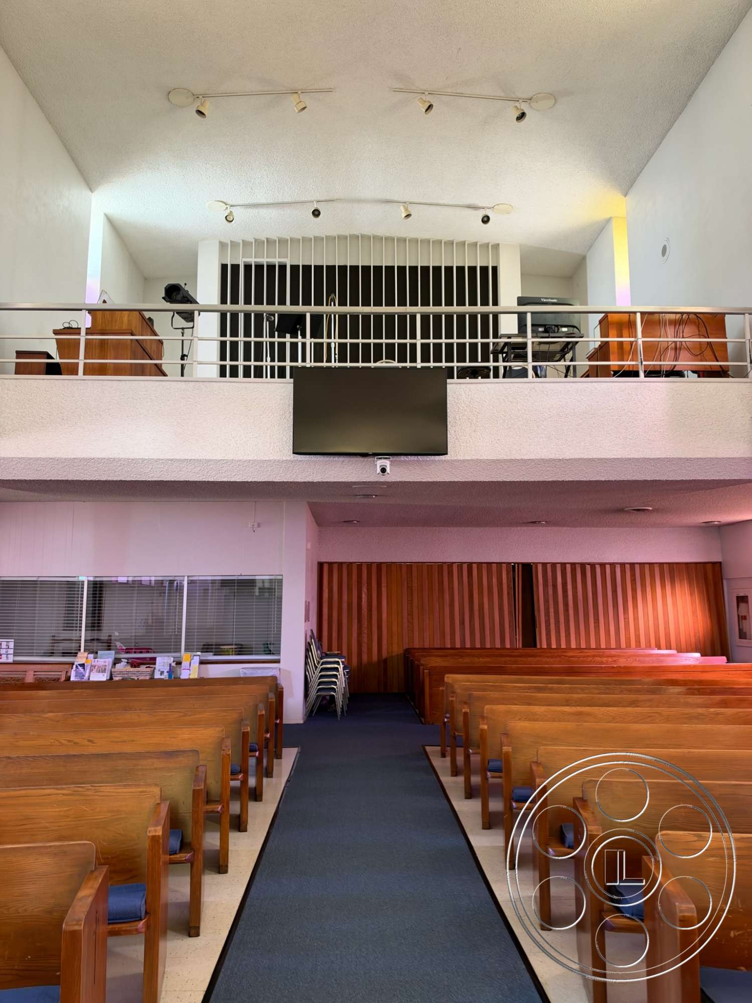 Community Center - wood panel accent wall, open floor layout, metal railing balcony, spotlight lighting fixtures, carpeted aisle, wooden pews with upholstered seats, exposed ceiling with textured finish, gray wall partitions, glass windows with horizontal slats, audio-visual equipment area, natural wood furnishings, formal worship space