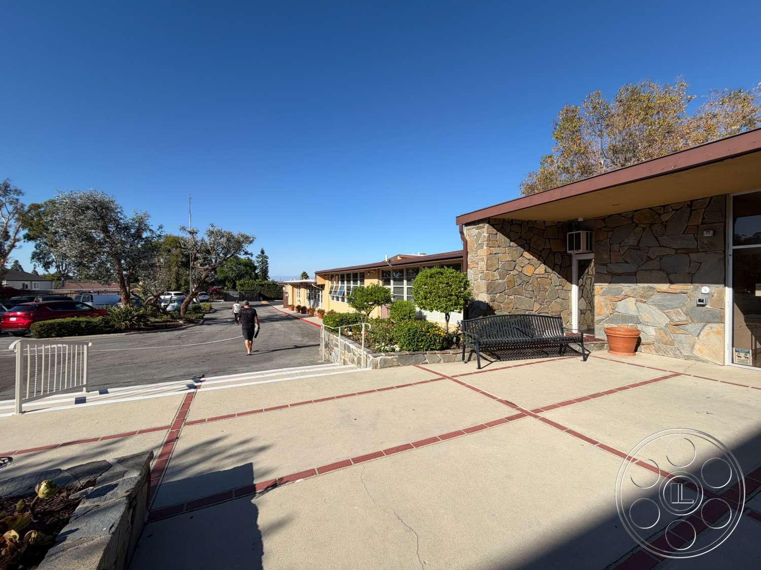 Community Center - exterior, landscaped garden, flat roof, potted plants, urban setting, modern architectural style, decorative pathway, single-story building, open parking area, concrete patio floor, stone façade wall, red brick inlay, metal park bench, entrance walkway