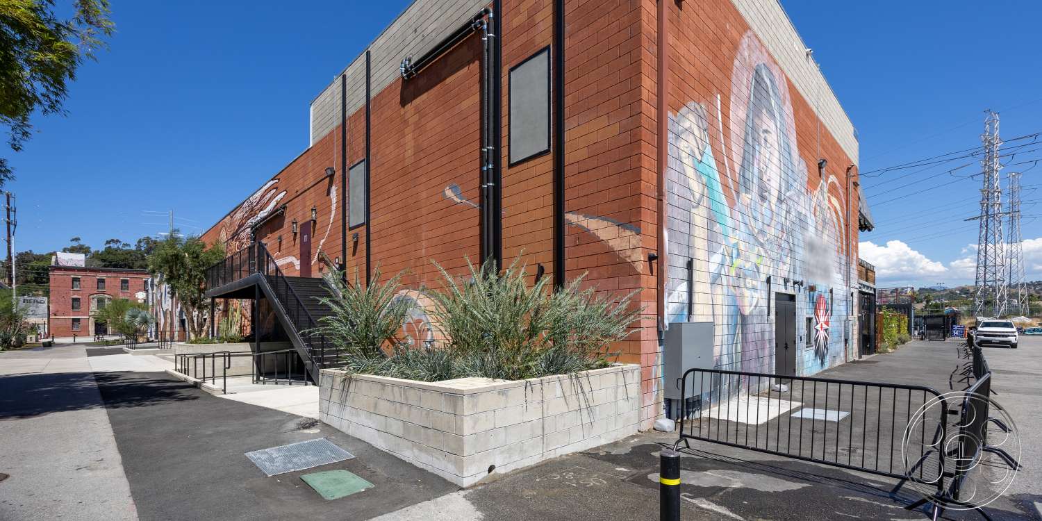 Club 74 - exterior, red brick walls, two-story building, modern industrial design, asphalt parking area, concrete planter boxes, metal railing stairs, mural artwork on brick, landscaped area with shrubs, utility pole with power lines