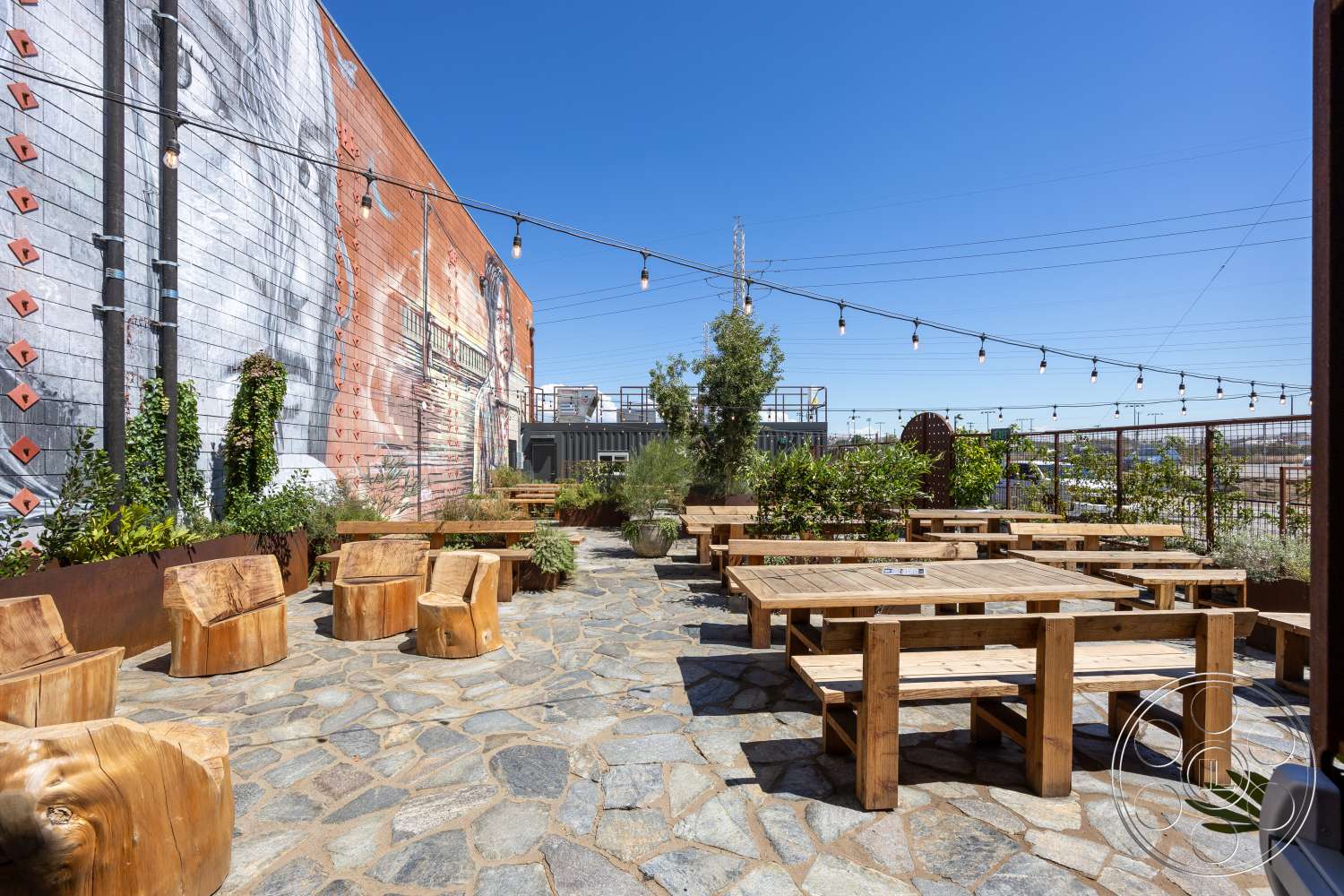 Club 74 - exterior, rustic planters, decorative string lights, urban outdoor space, stone patio flooring, lush greenery landscaping, metal fence railings, open sky setting, wood picnic tables, wood stump seating, mural wall art, café-style seating area