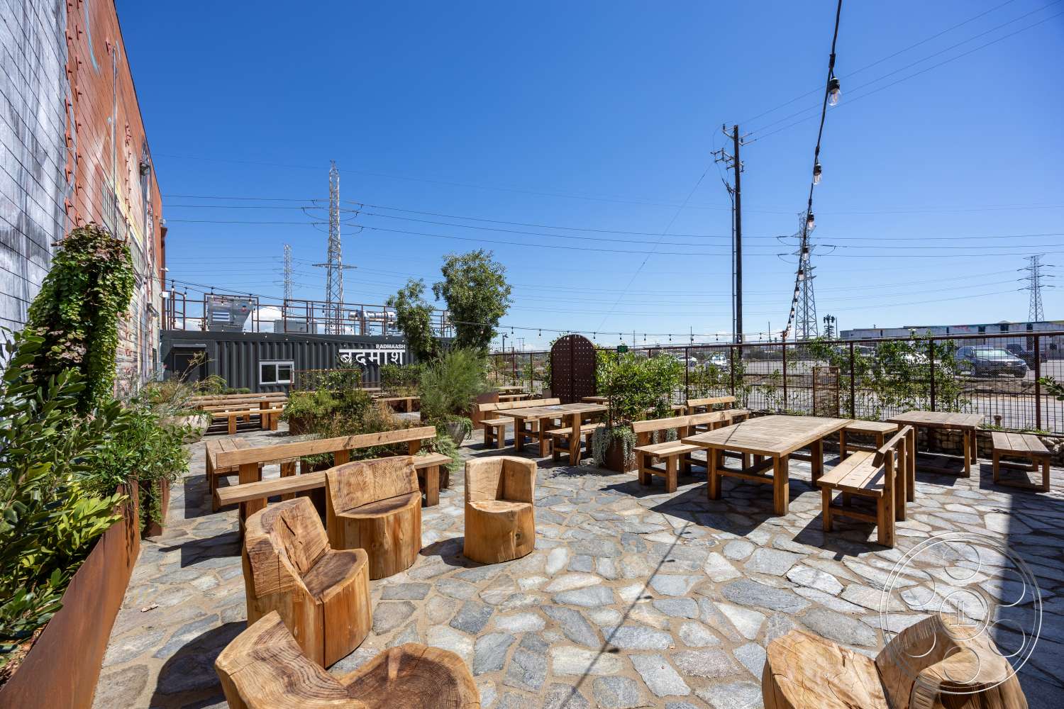 Club 74 - exterior, green landscaping, metal fence, clear blue sky, wooden seating, rustic wooden chairs, stone patio flooring, urban outdoor setting, wood outdoor dining tables, industrial backdrop, decorative wooden archway