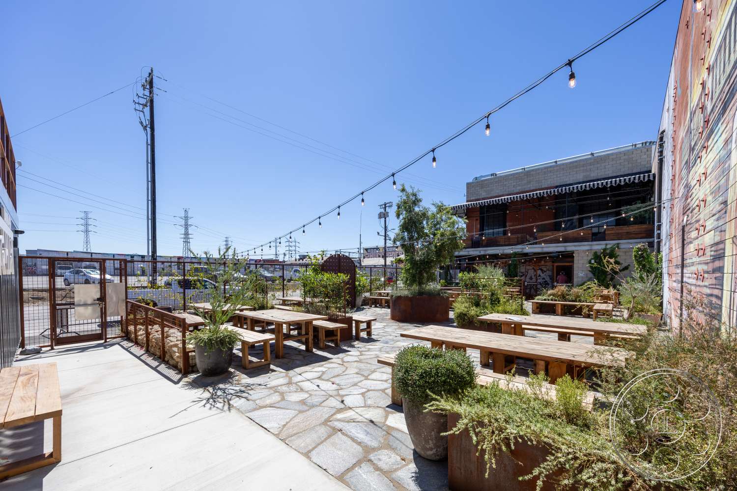 Club 74 - exterior, outdoor seating area, urban setting, planted greenery, metal fencing, wooden picnic tables, concrete walkways, string lighting, stone patio flooring, industrial-style building, rust-colored metal planters, decorative stone paths
