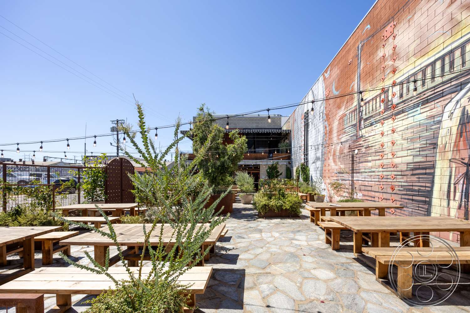 Club 74 - exterior, hanging string lights, wooden picnic tables, contemporary urban setting, stone patio flooring, open outdoor seating area, mural wall art, decorative metal fencing, greenery and plants landscaping