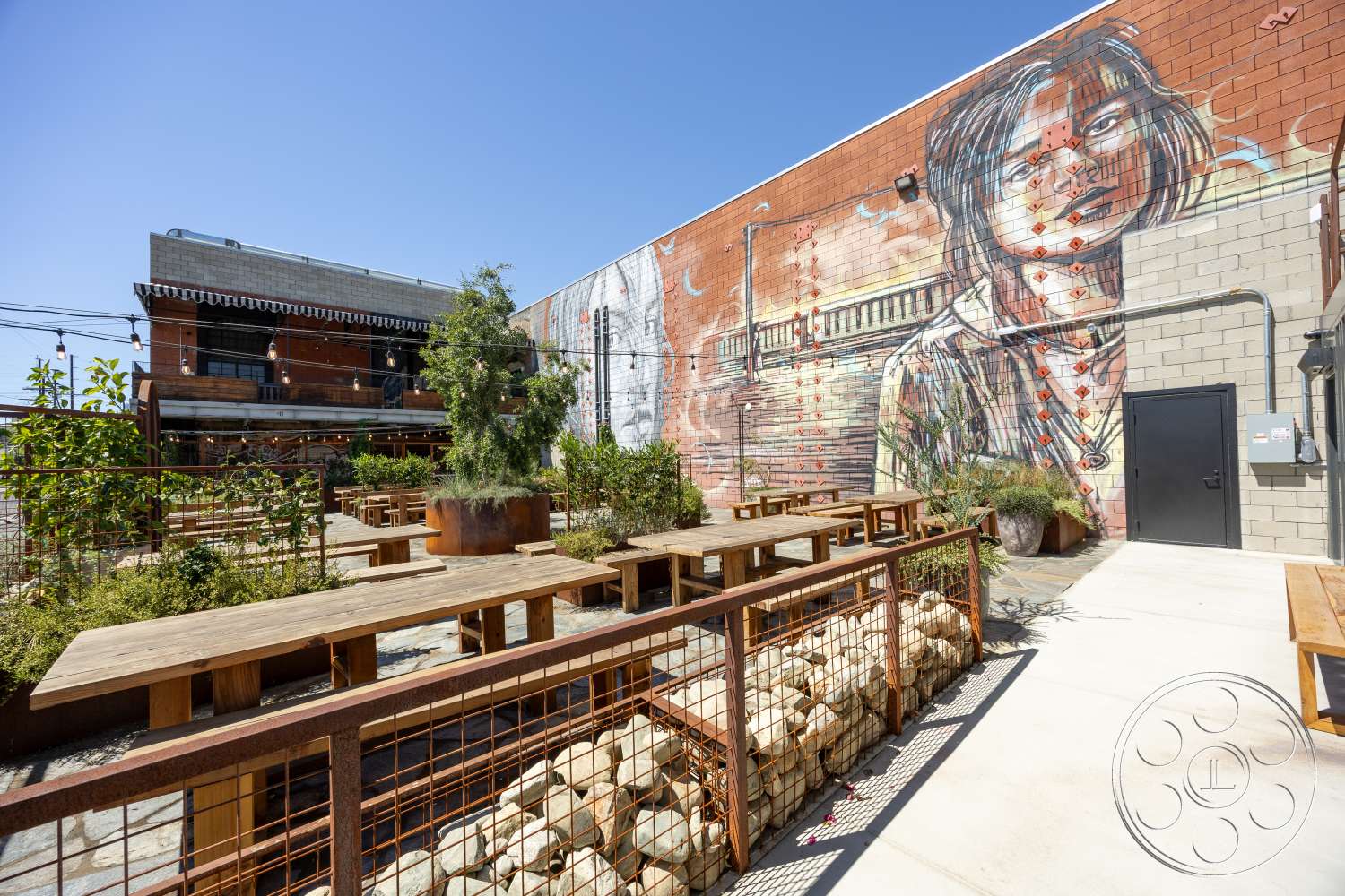 Club 74 - exterior, wooden picnic tables, concrete patio flooring, exposed concrete columns, overhead string lights, urban outdoor seating area, industrial-style building, brick wall with mural, rustic metal fencing, steel wire mesh planters, landscaped garden with plants, decorative gravel landscape