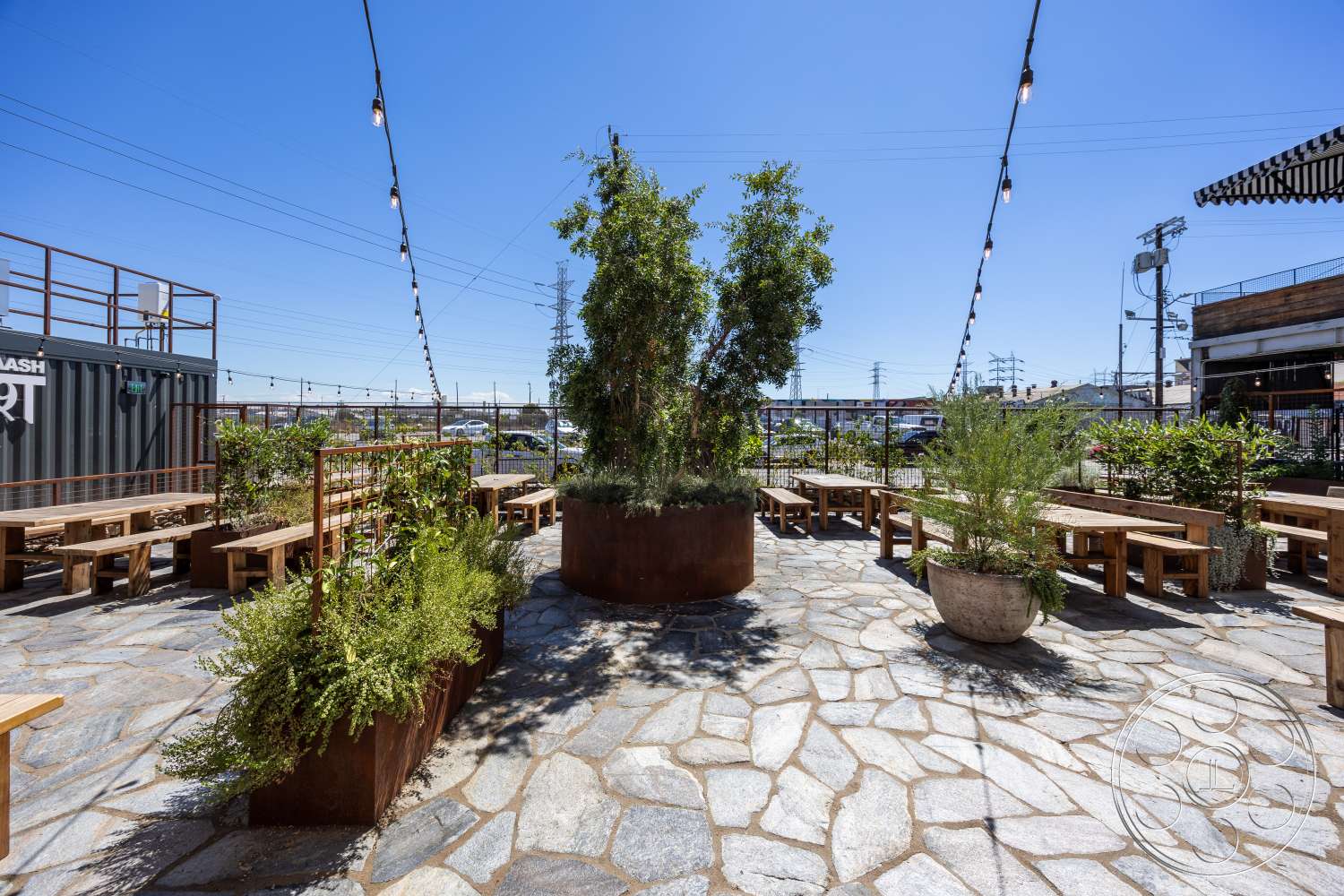Club 74 - exterior, urban environment, sunny outdoor setting, flagstone patio flooring, string outdoor lighting, green foliage landscaping, decorative stone feature wall, rust-colored metal planters, rustic wooden picnic tables, industrial container design, open air seating area