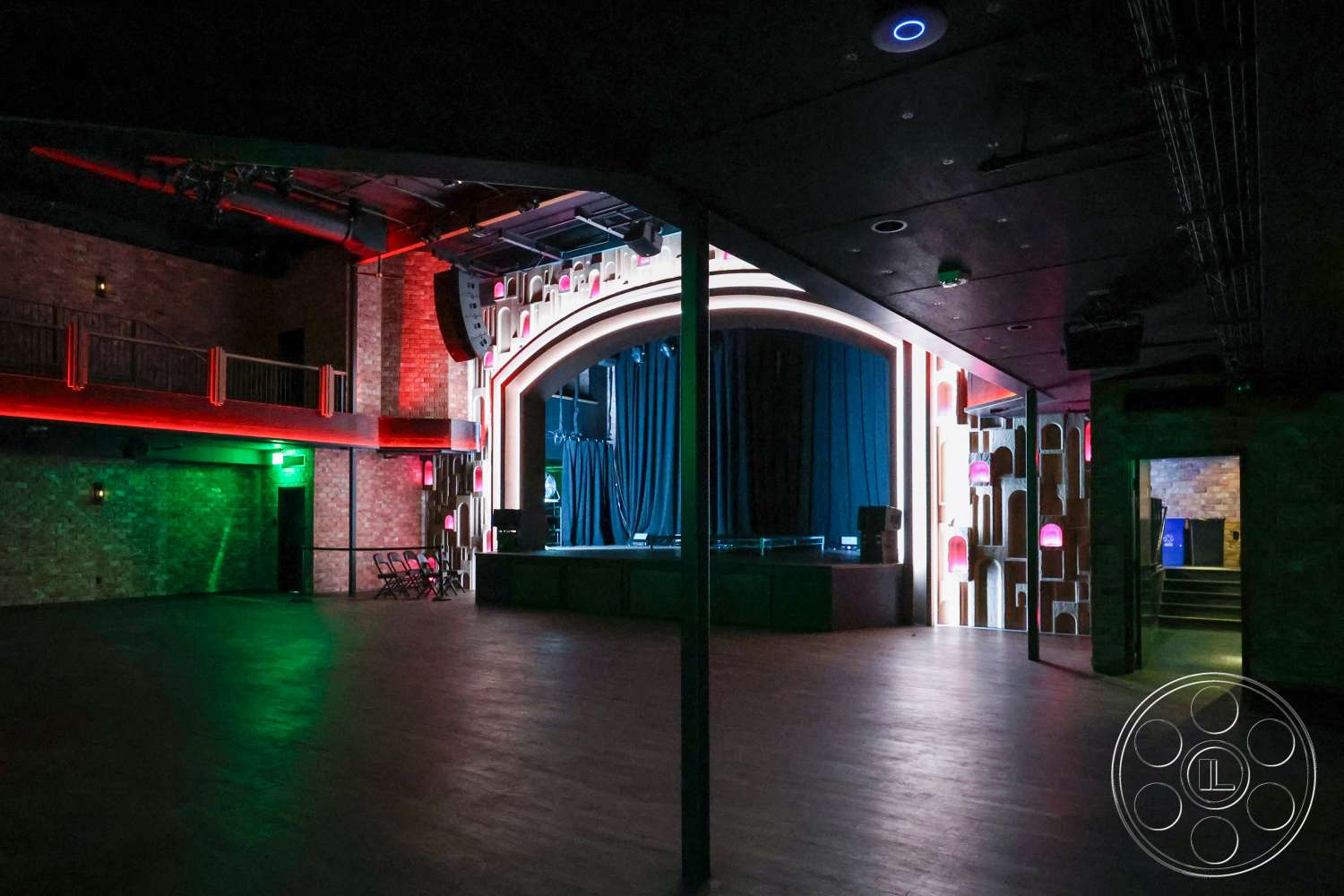 Club 74 - exposed brick walls, industrial lighting fixtures, black hardwood flooring, steel support beams, open performance space, tiered seating area, arched stage backdrop, stage with blue curtain