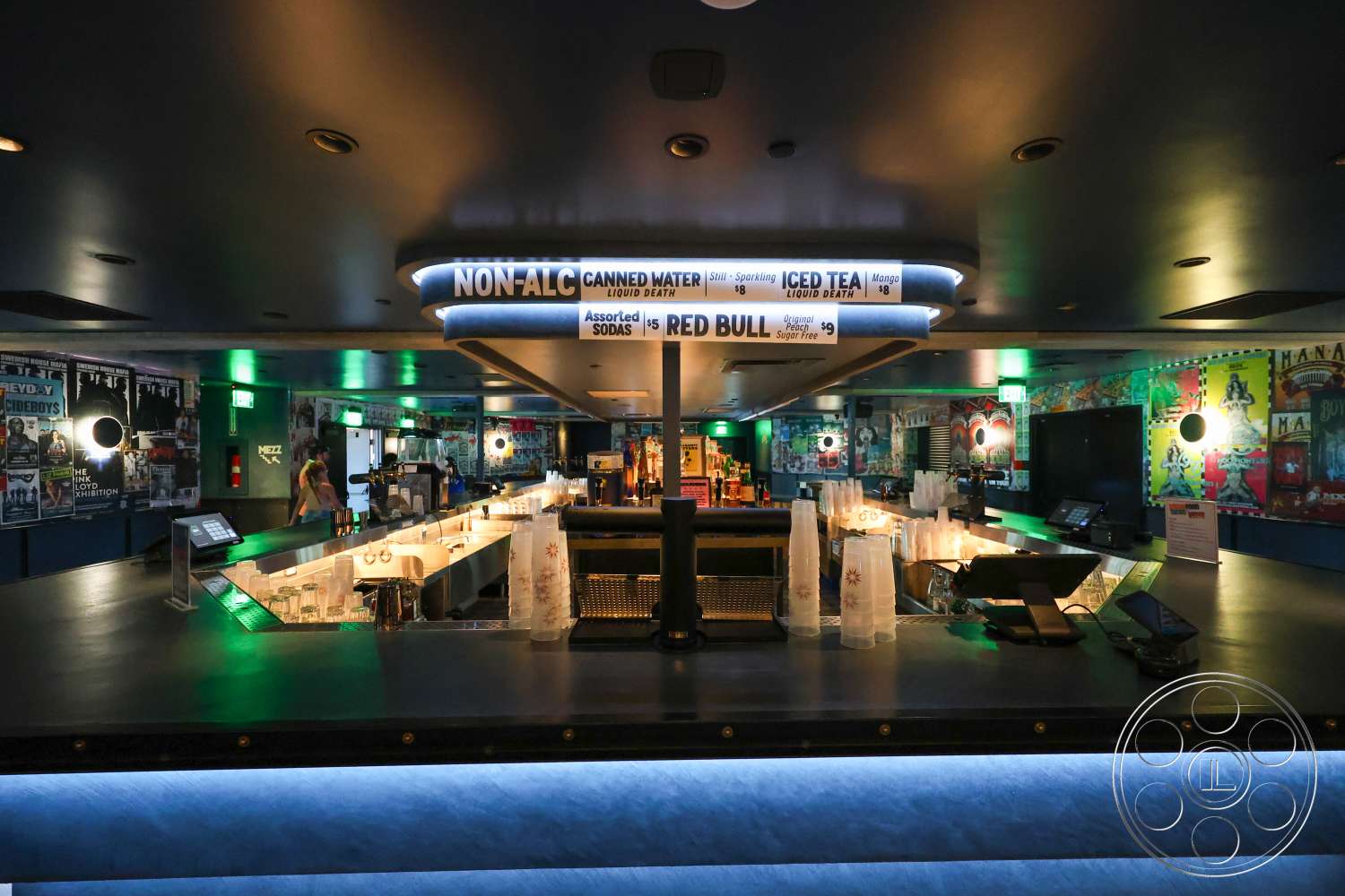 Club 74 - black countertop, modern bar stools, wood paneling accents, industrial style lighting fixtures, interior bar area, dark ceiling with recessed lighting, illuminated bar back, metallic bar shelving, framed posters on walls, glassware arrangement on counter, LED strip lighting under bar, signage with menu items, cocktail preparation station