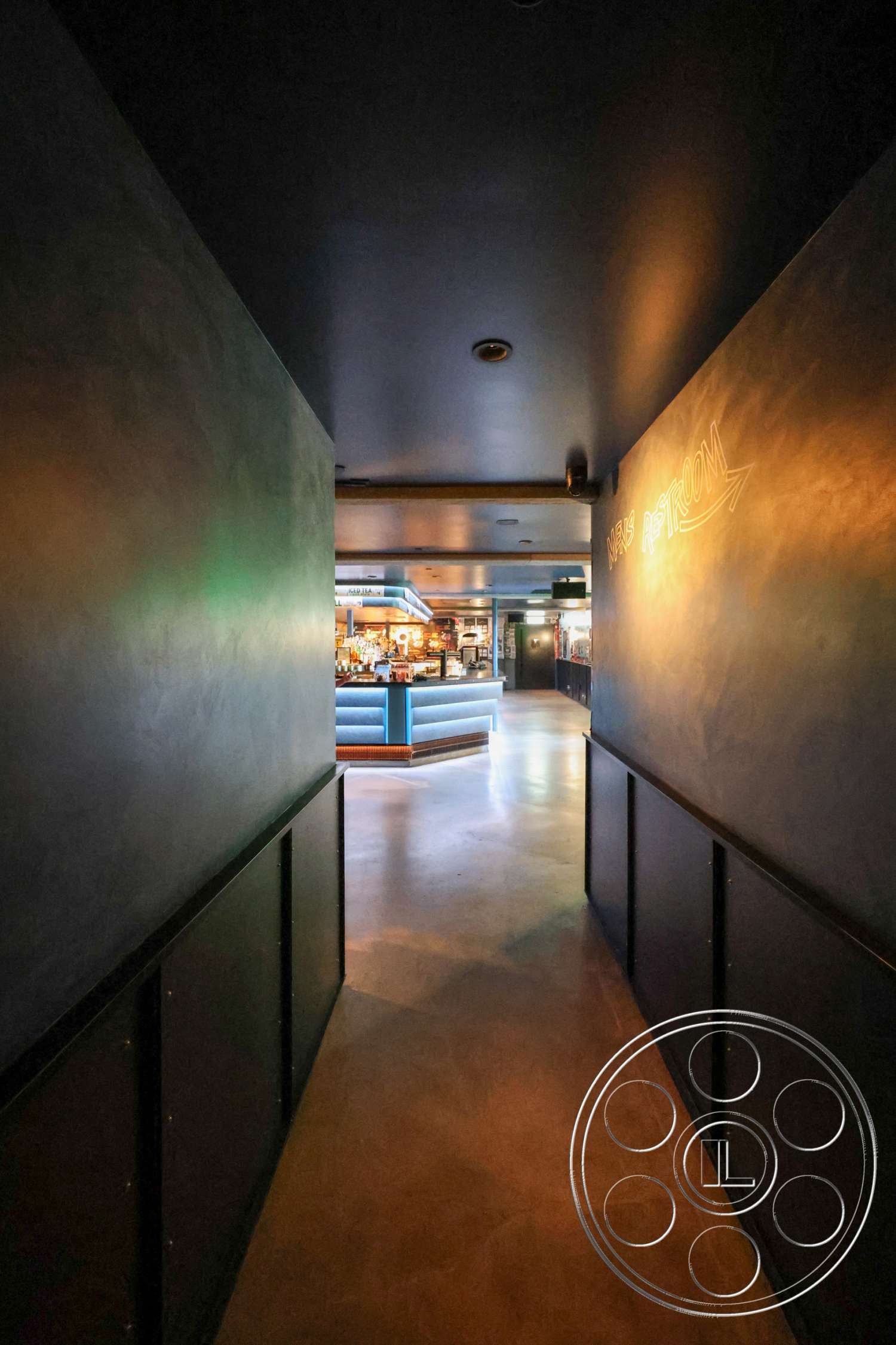 Club 74 - polished concrete flooring, black wood paneling, interior hallway, sleek modern design, ceiling-mounted lighting fixtures, dark plaster walls, dimly lit walkway, restaurant bar area view, blue illuminated bar counter